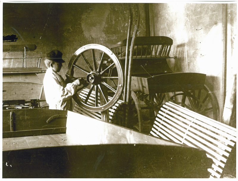Svartvit bild på ett barn med ett vagnshjul i en träverkstad.