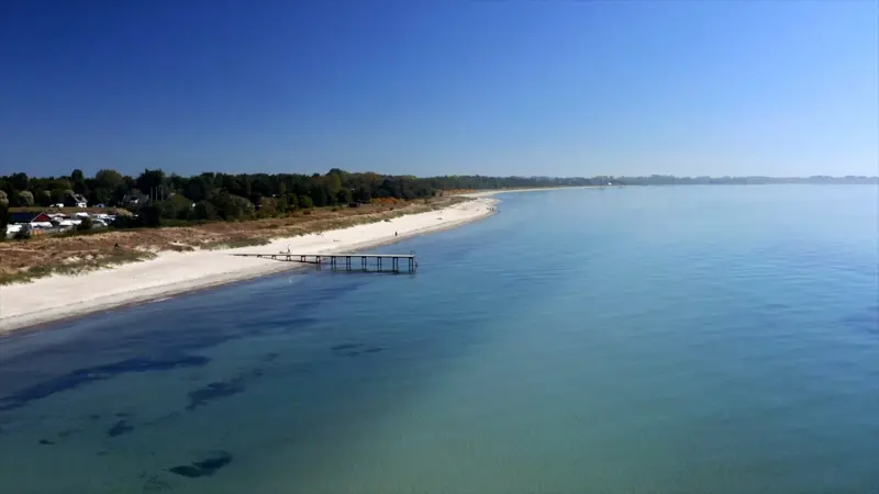 En kuststräcka fortograferad uppifrån. Vackert blågrönt vatten och en sandstrand med en brygga