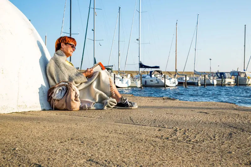 En person sitter och läser i en hamn, solsken och blå himmel, båtar i bakgrunden.