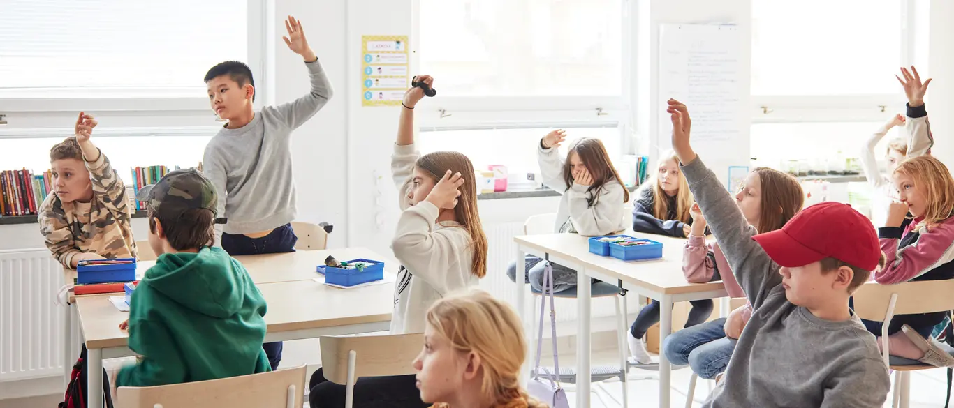 Barn i ett klassrum. En del räcker upp handen. Det är ljust och stämningen känns levande. 