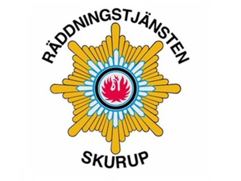Ett guldemblem med snirklar runt en röd rovfågelliknande figur. Kring emblemet står det Räddningstjänsten Skurup.