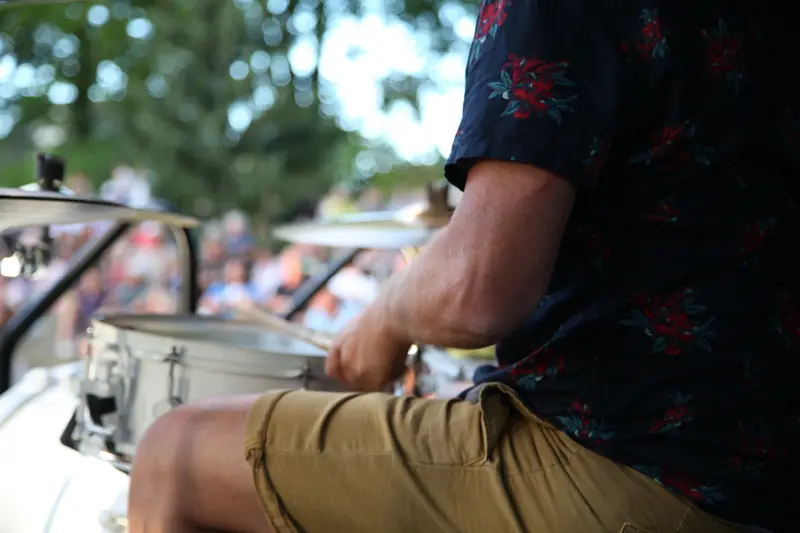 A drummer is seen from behind/the side. We see part of his thigh and a drum, one of his arms and a drum stick. 