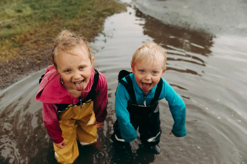 två mindre barn i regnkläder leker och busar i vattenpöl